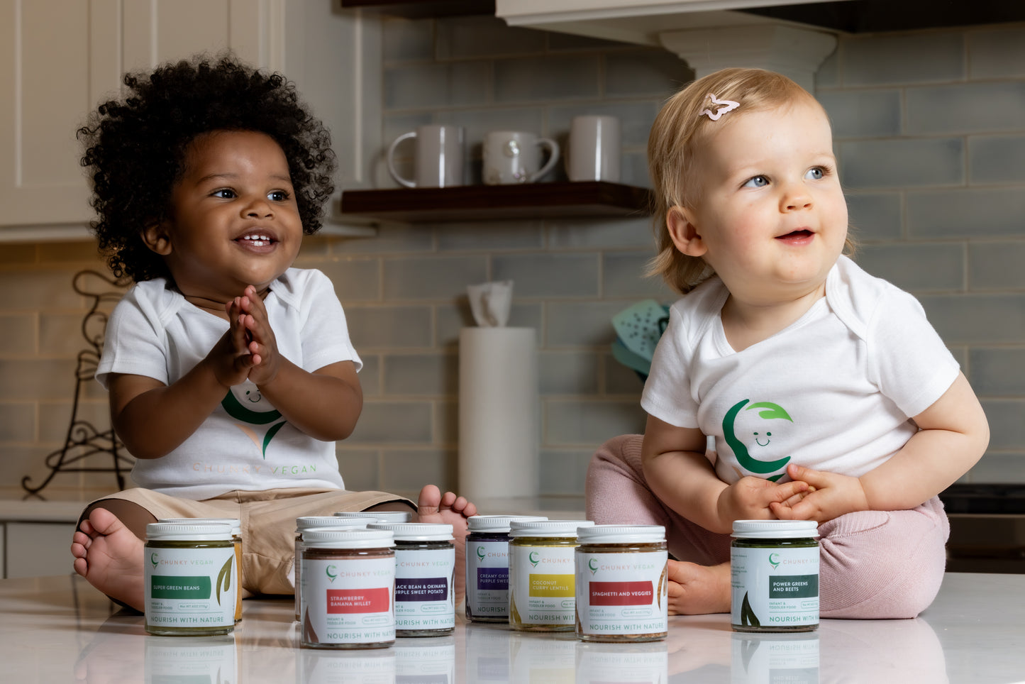 Two children sitting on a kitchen counter with jars of food products in front of them.