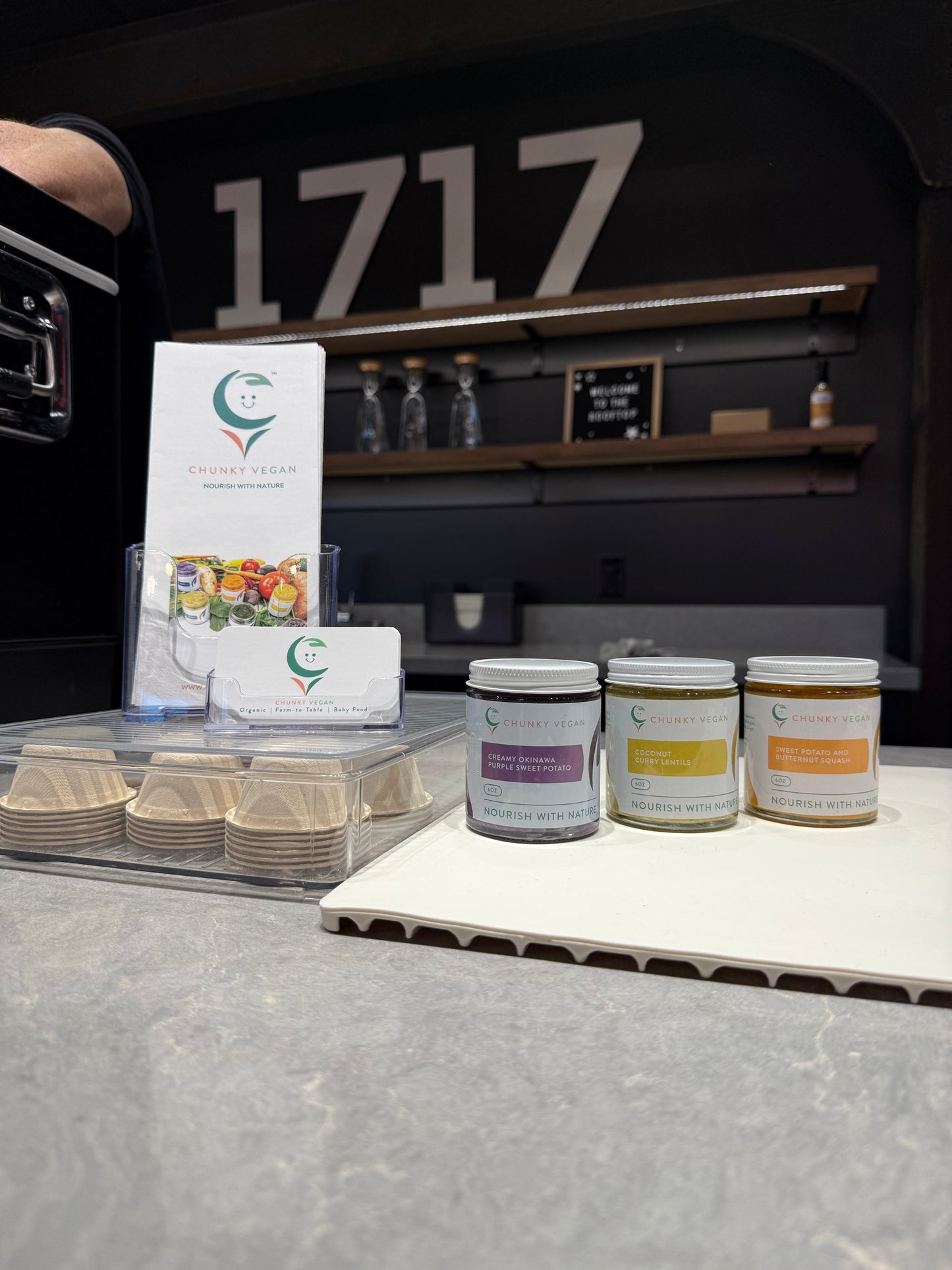 Three jars with labels on a table in front of a display stand with brochures.
