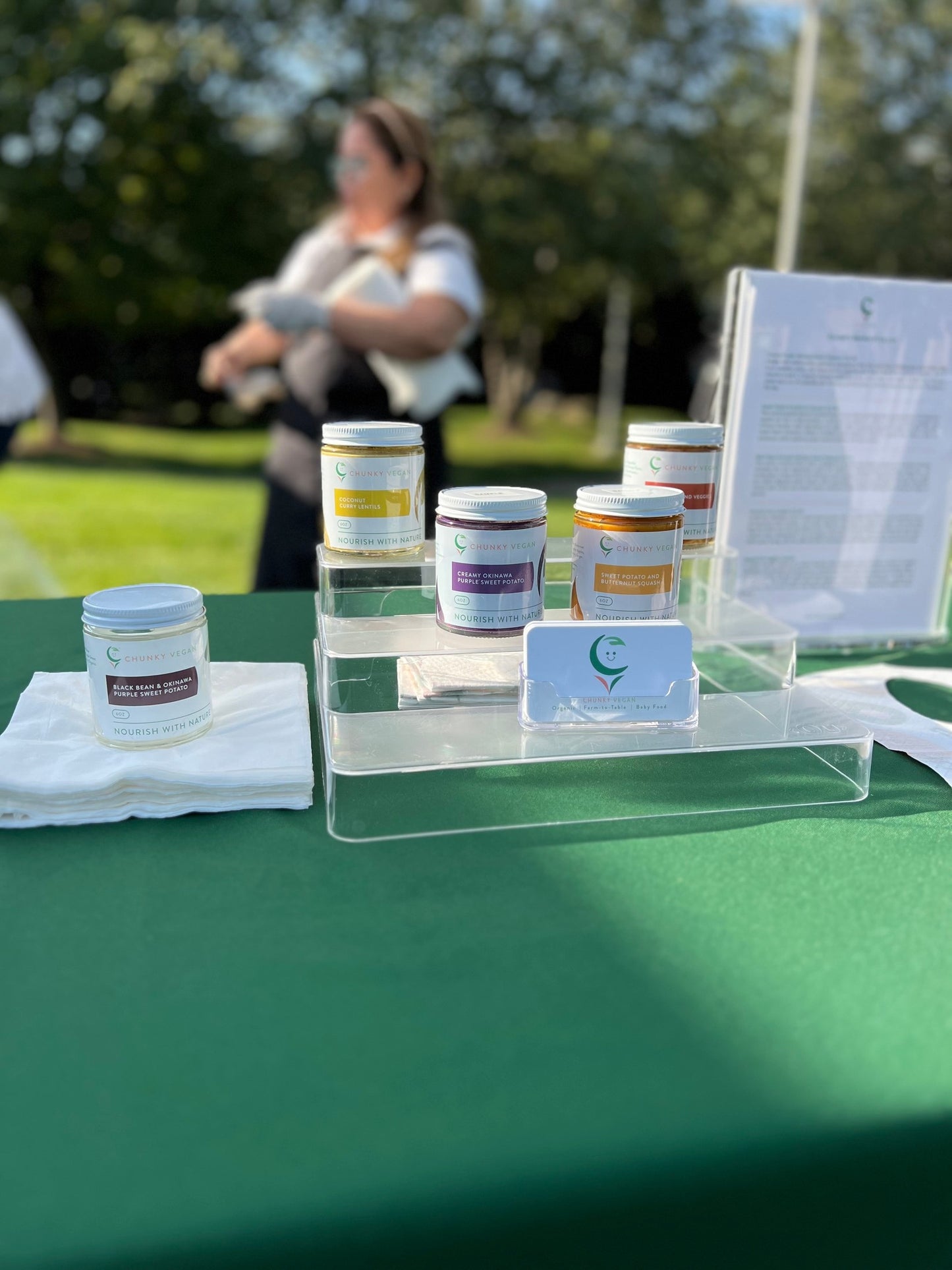 Small containers of Chunky Vegan Baby Food on a table with a blurred background of people and greenery