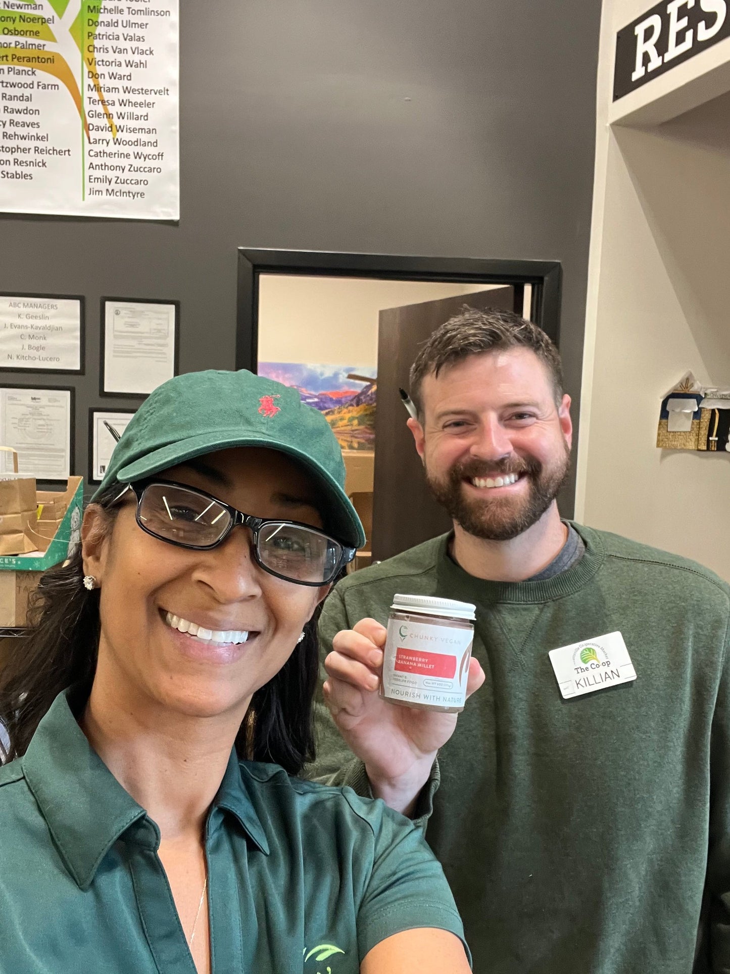 Two people in green shirts holding a jar of Chunky Vegan Baby Food, with a wall of framed documents in the background at Lovettesville Co-Op
