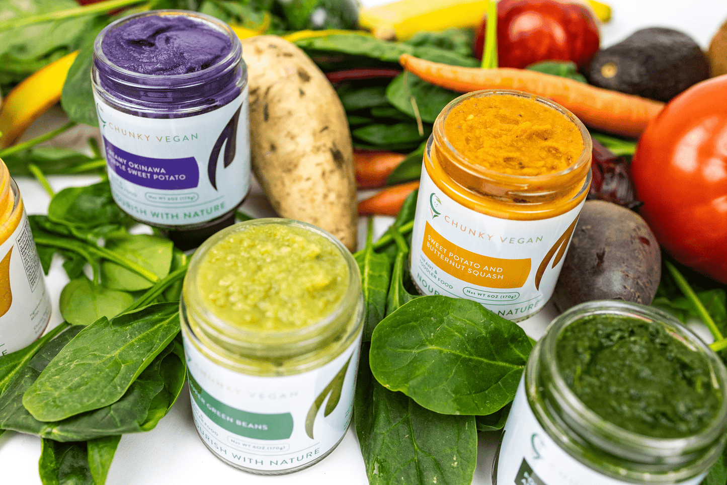 Jars of baby food surrounded by fresh vegetables on a white background