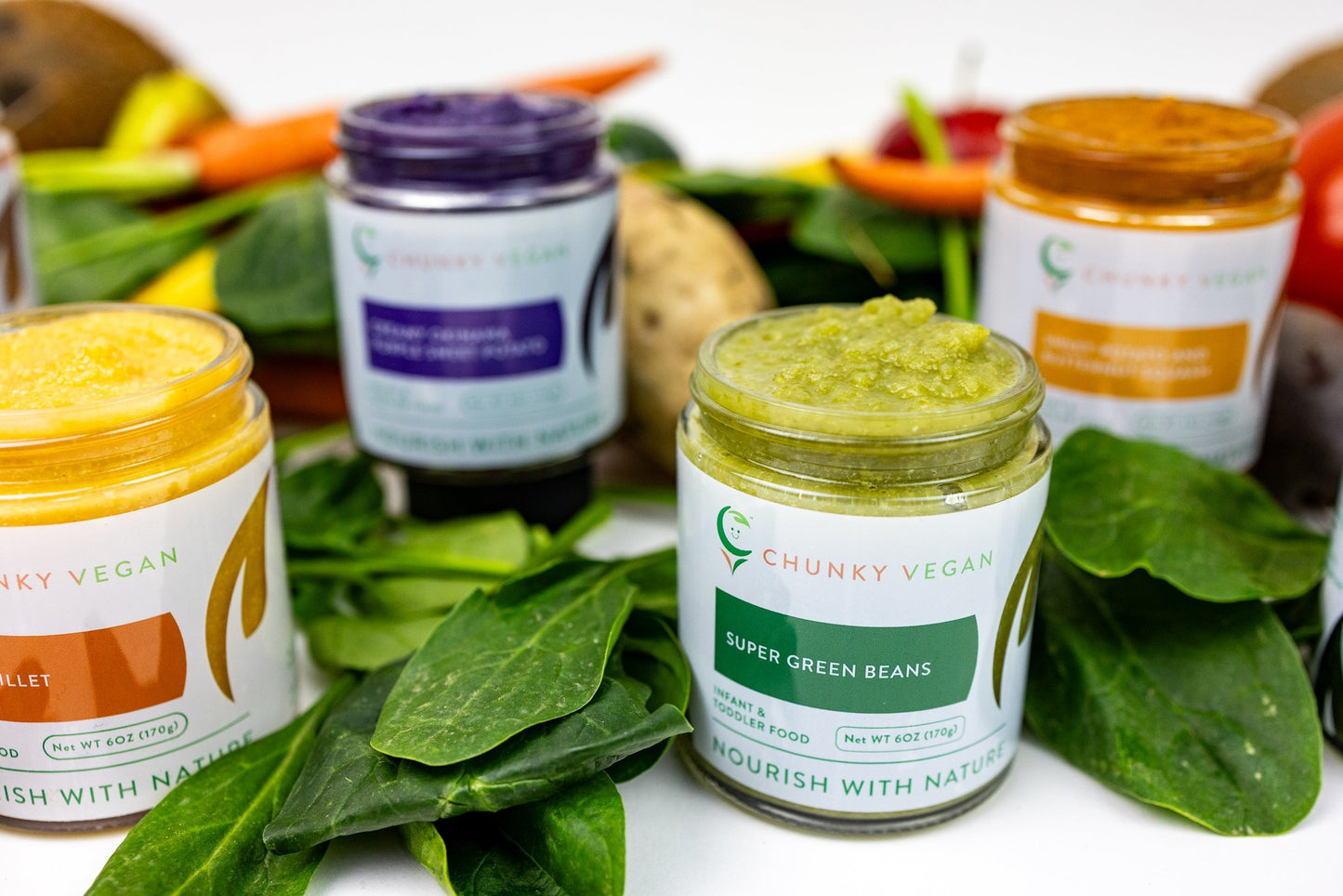 Jars of Chunky Vegan food products surrounded by fresh vegetables on a white background